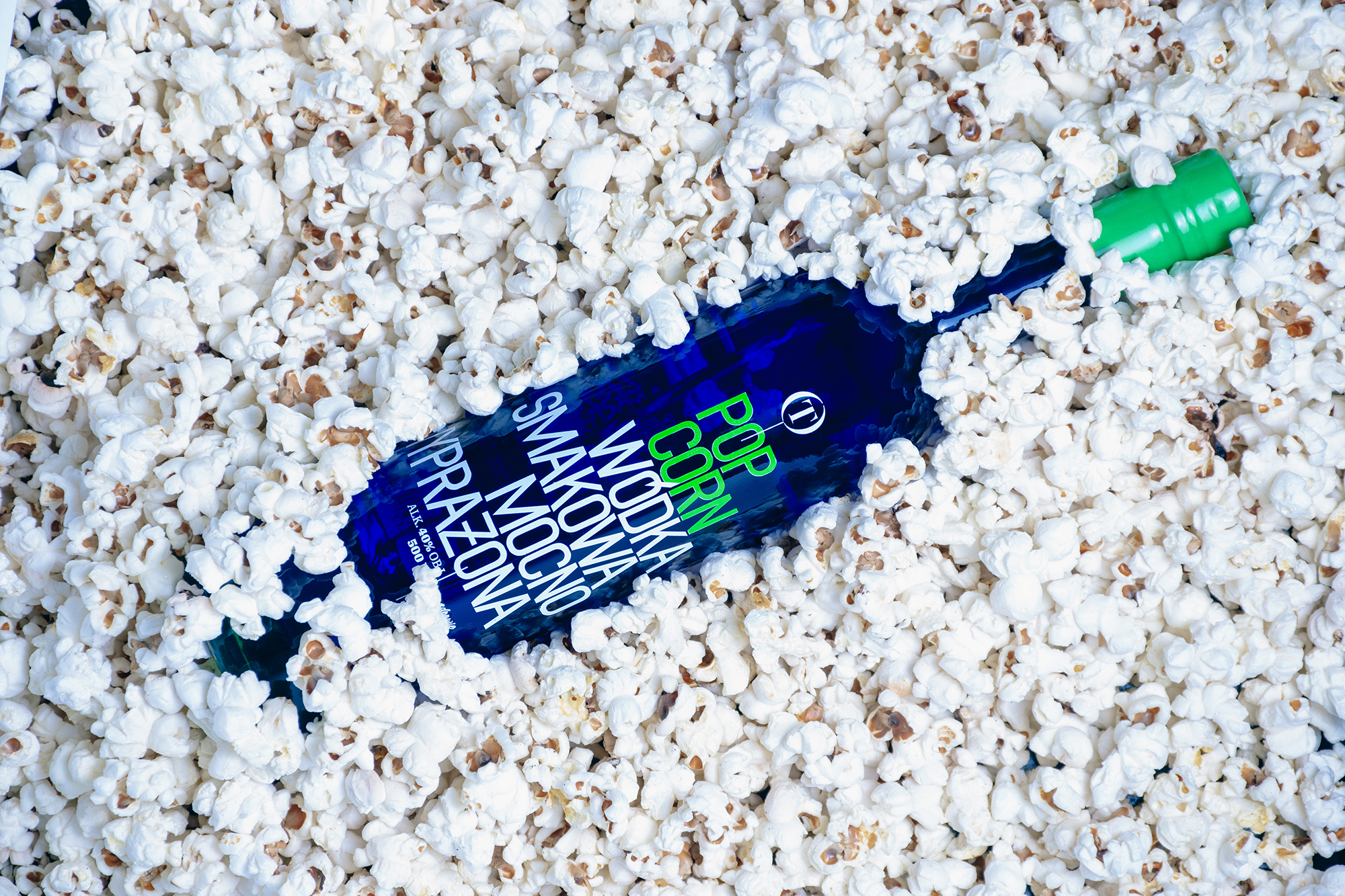 Niebieska butelka wódki smakowej popcorn otoczona białym popcornem.
