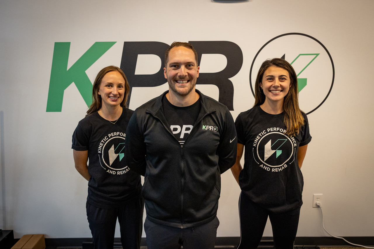 Three people smiling, wearing black shirts with Kinetic Performance and Rehab logo standing indoors.