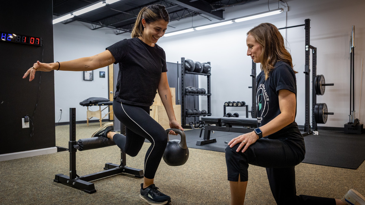 Woman performing weighted lunge exercise in gym while another woman watches and coaches her.