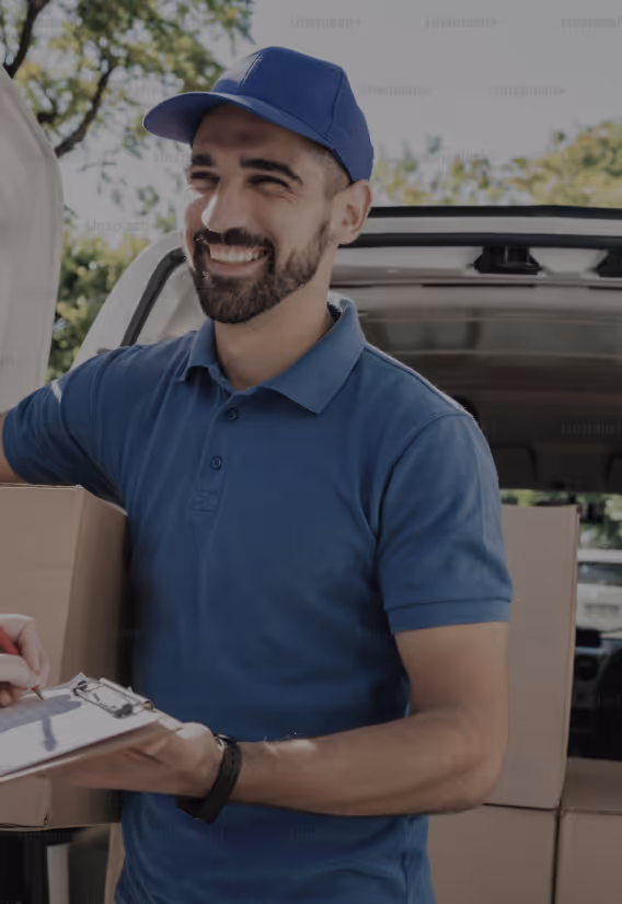 Hymyilevä jakelija sinisessä univormussa seisoo pakettiauton vieressä pakattujen laatikoiden kanssa ja pitää leikepöytää.