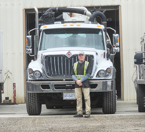 Man in safety vest standing in front of a large white industrial truck outside Economy Vacuum Tankers & Toilet Rentals.