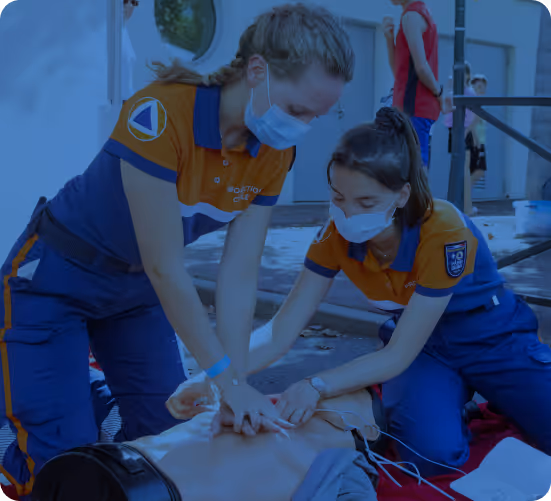 Two emergency responders in orange and blue uniforms performing CPR on a training mannequin outdoors, both wearing face masks.