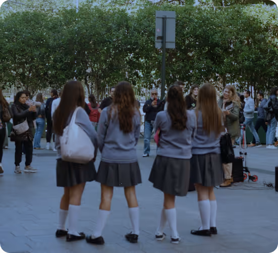 Four schoolgirls in grey sweaters, skirts, and white knee-high socks standing in an outdoor area with people and trees in the background.