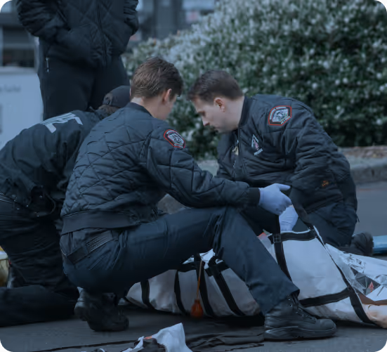Three emergency responders in dark jackets attending to a person lying on the ground wrapped in a white body bag.