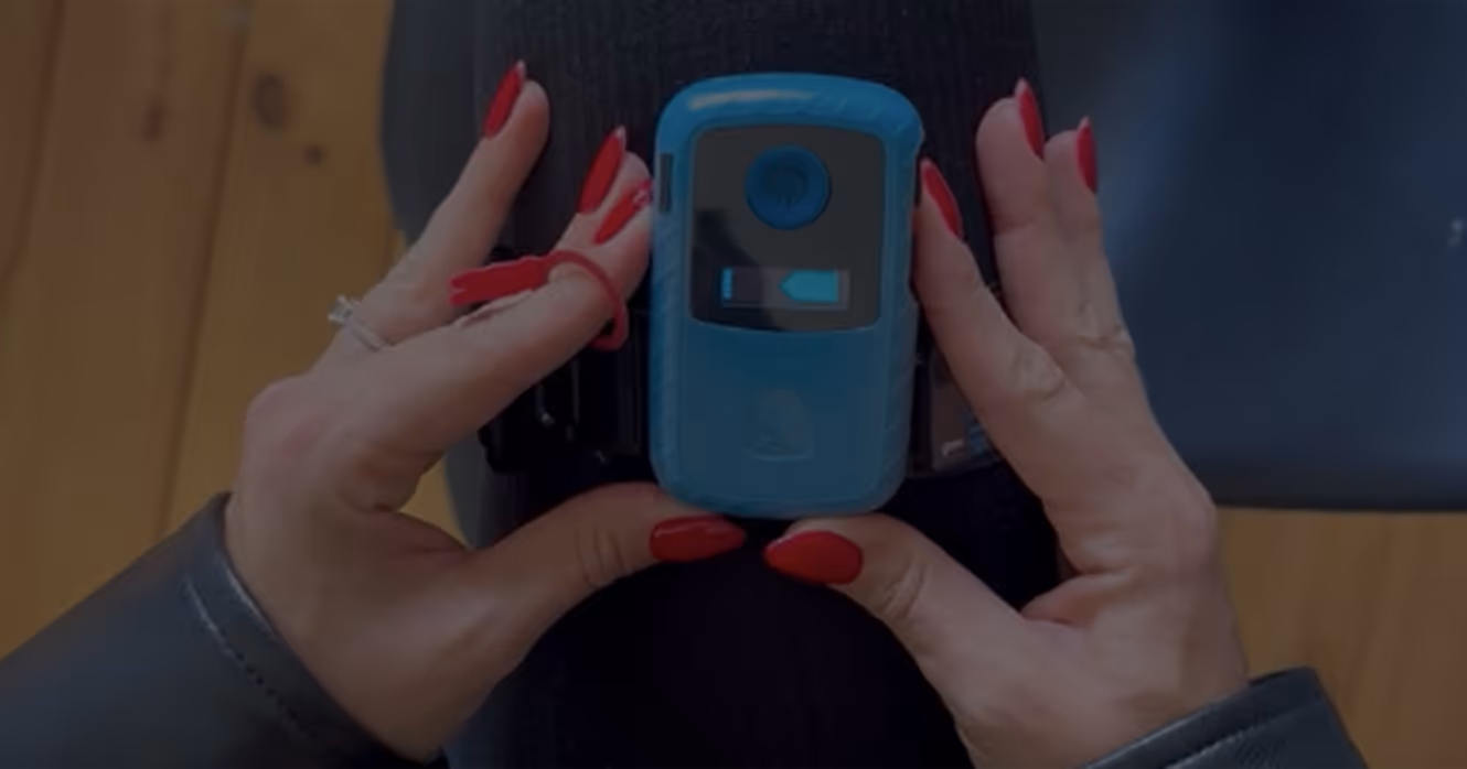 Hands with red nail polish holding a blue device with a screen and power button, strapped around a black fabric surface.