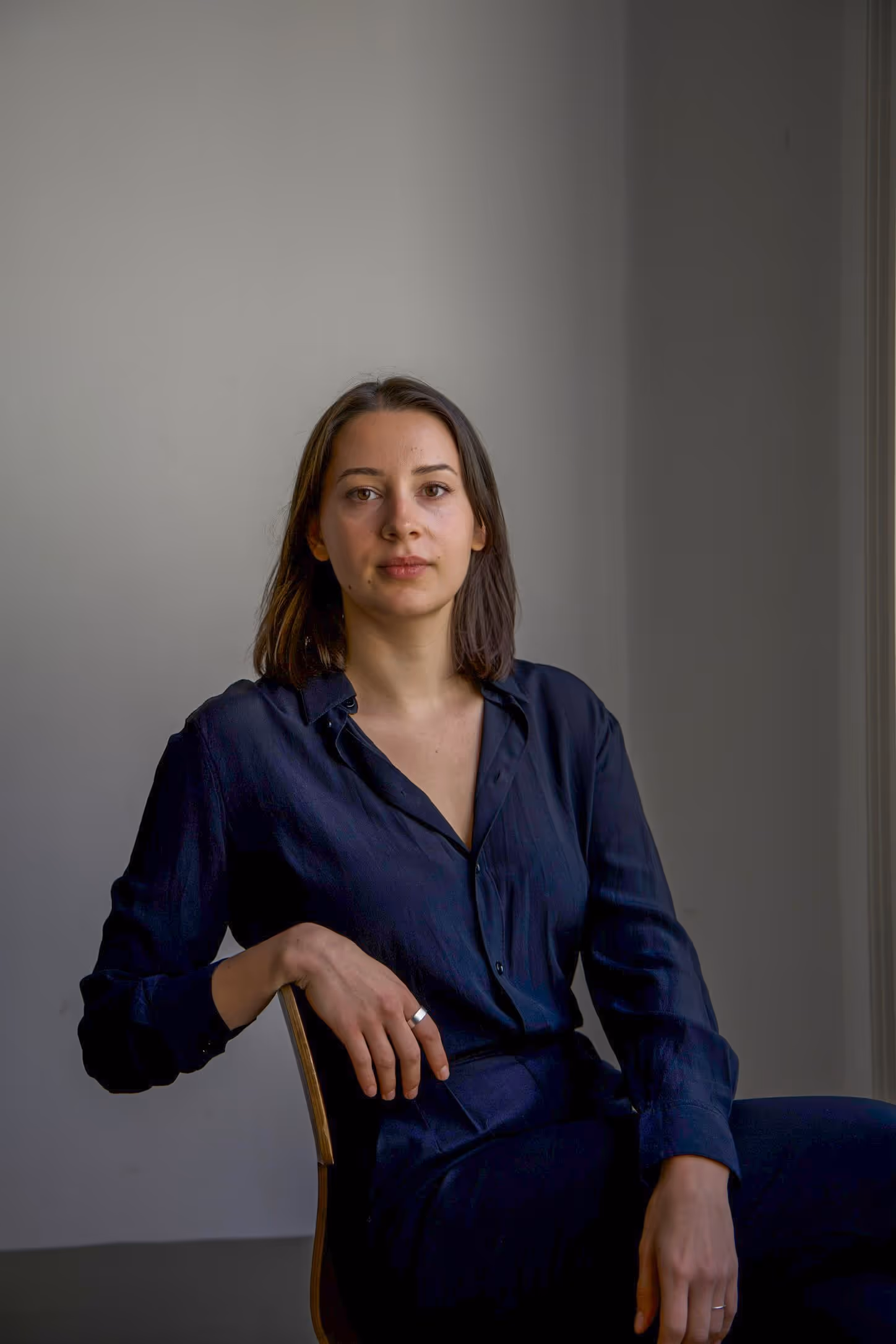 Woman with shoulder-length dark hair wearing a dark navy button-up shirt sitting on a chair against a plain gray background.