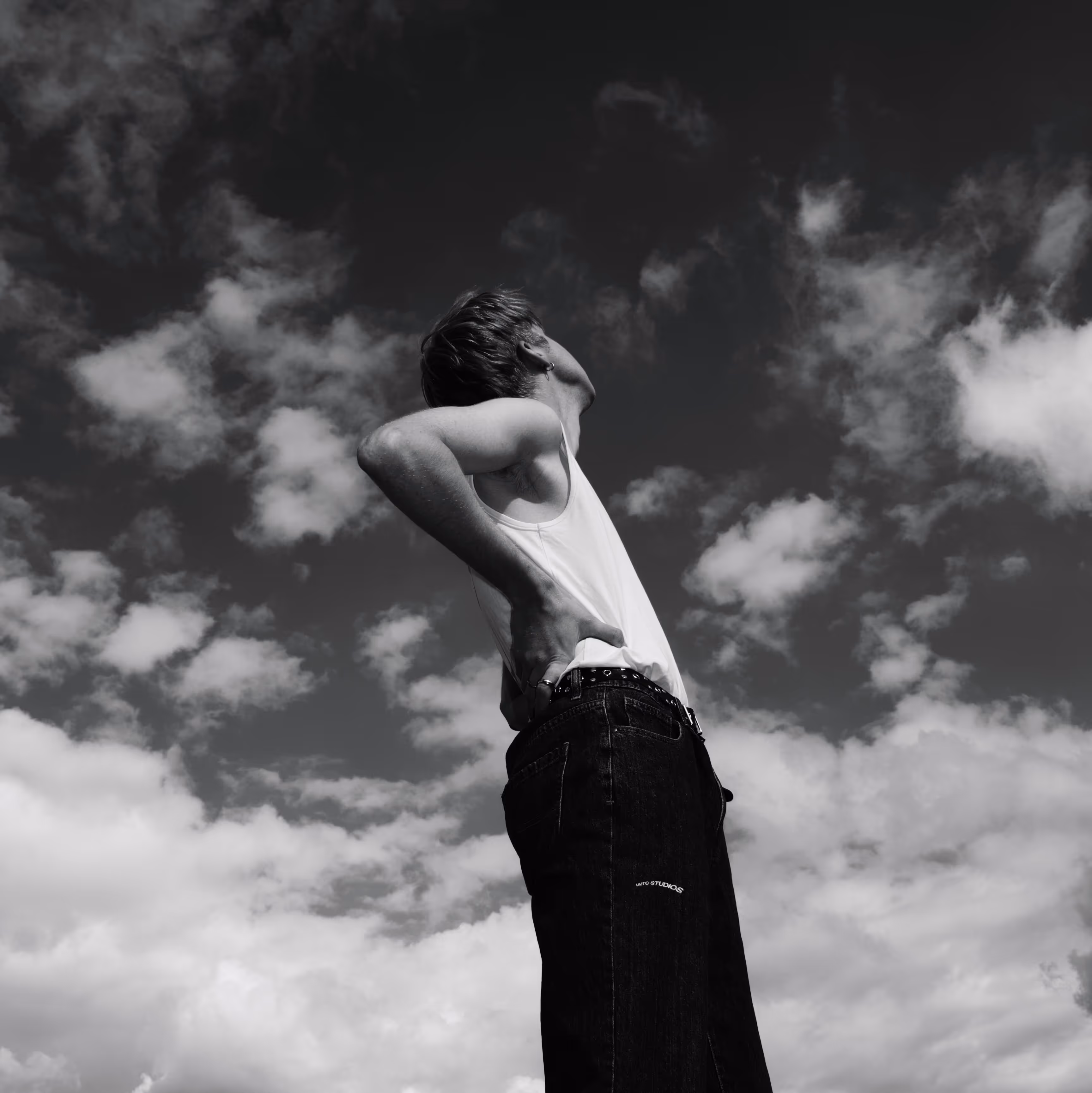 Black and white photo of a person in a white sleeveless top and dark pants standing with hands on hips, looking upward against a cloudy sky.