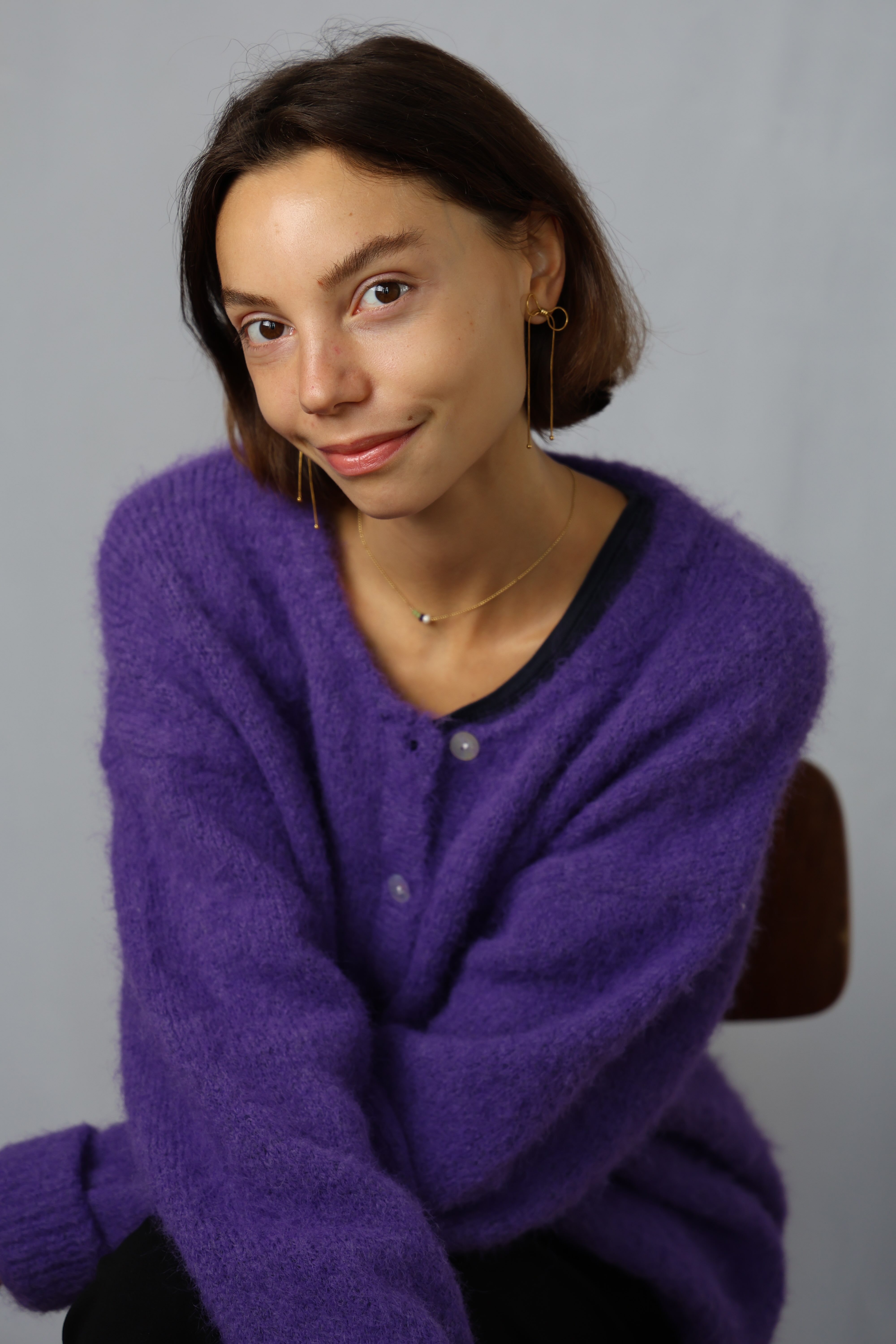 Young woman with short brown hair and gold earrings wearing a purple fuzzy cardigan, smiling softly against a plain gray background.