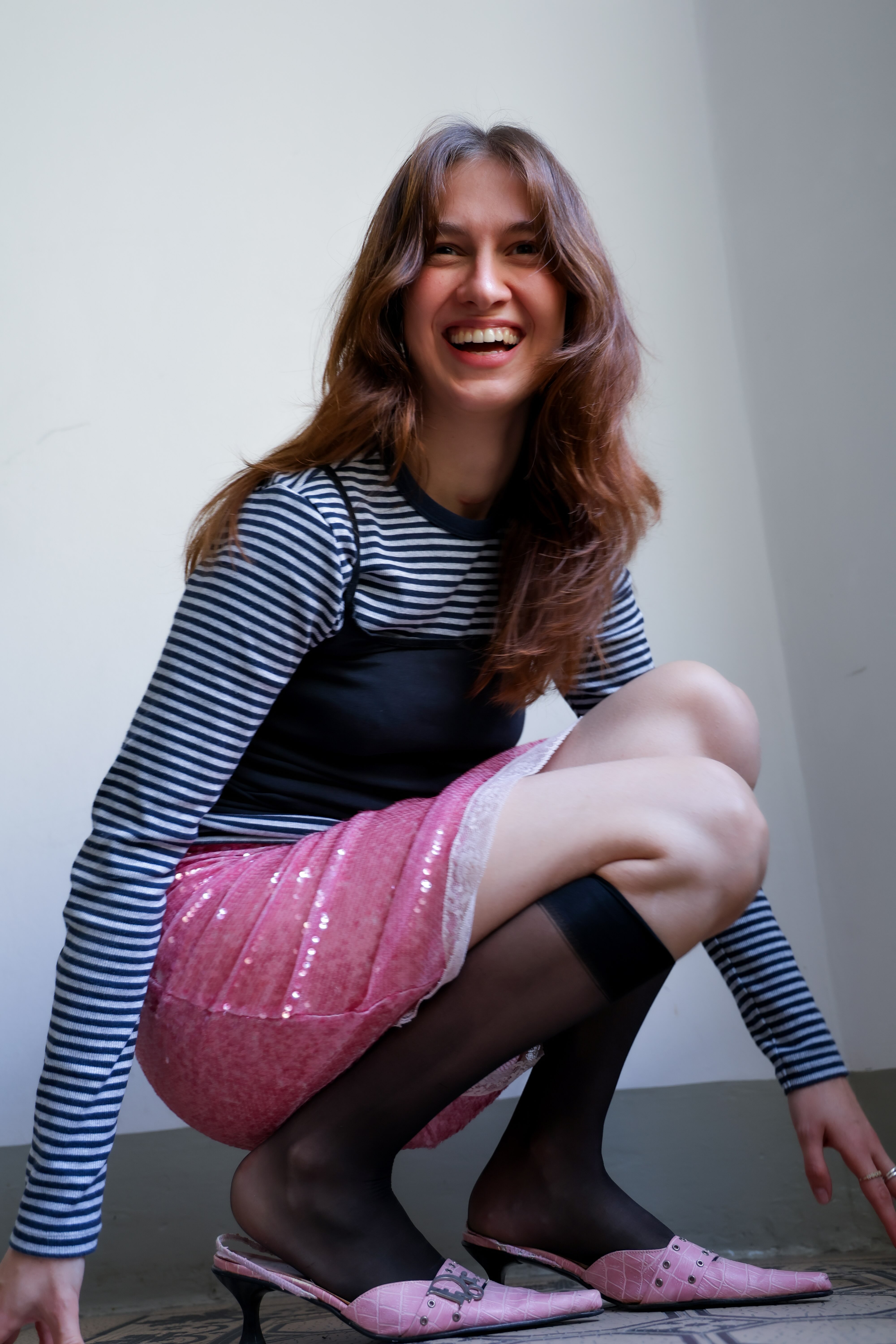Smiling woman with long brown hair squatting indoors wearing a striped long-sleeve shirt, pink skirt, black stockings, and pink heeled shoes.