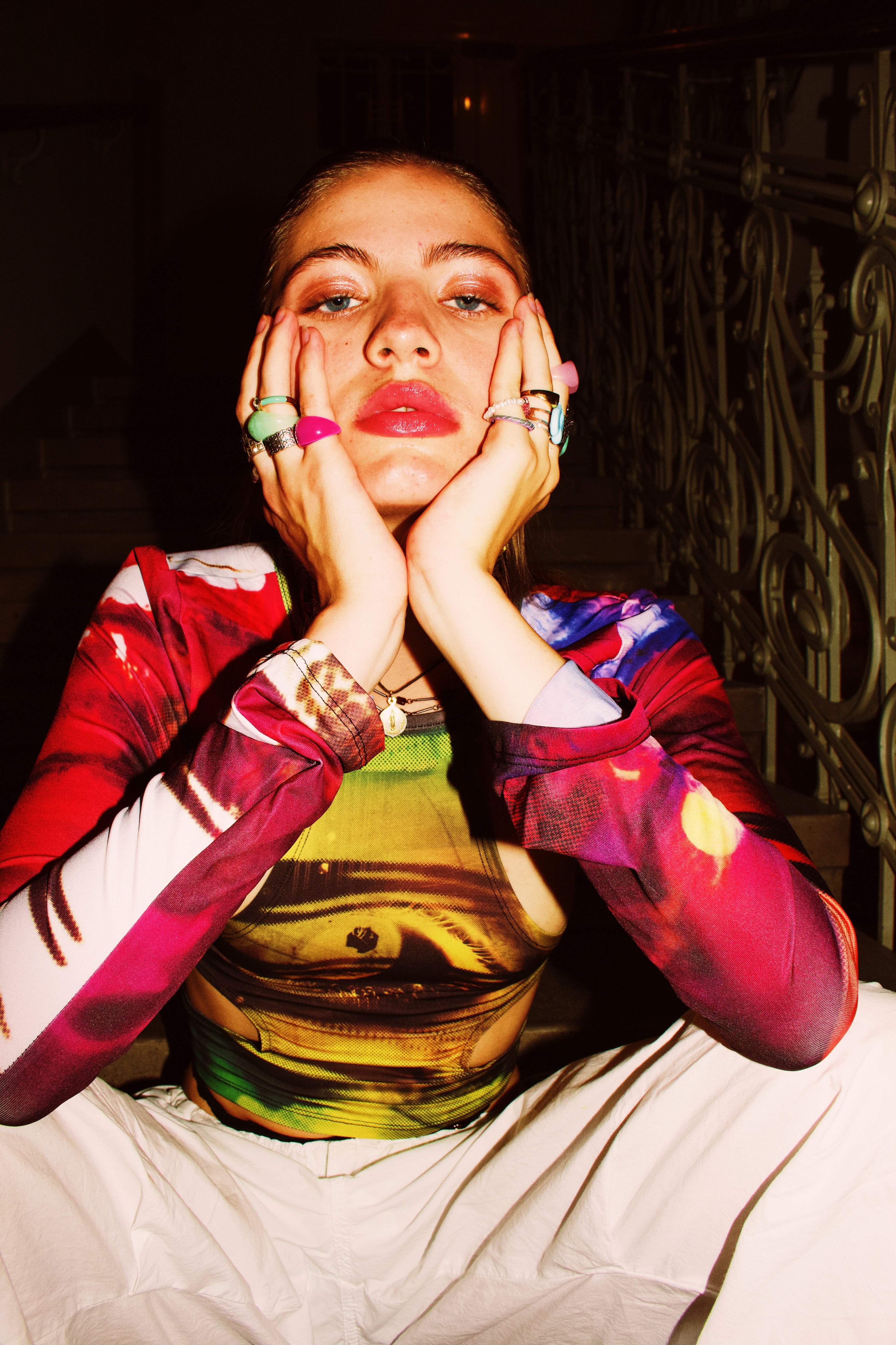 Young woman with colorful rings and vibrant top resting her face in her hands, sitting indoors by a staircase.