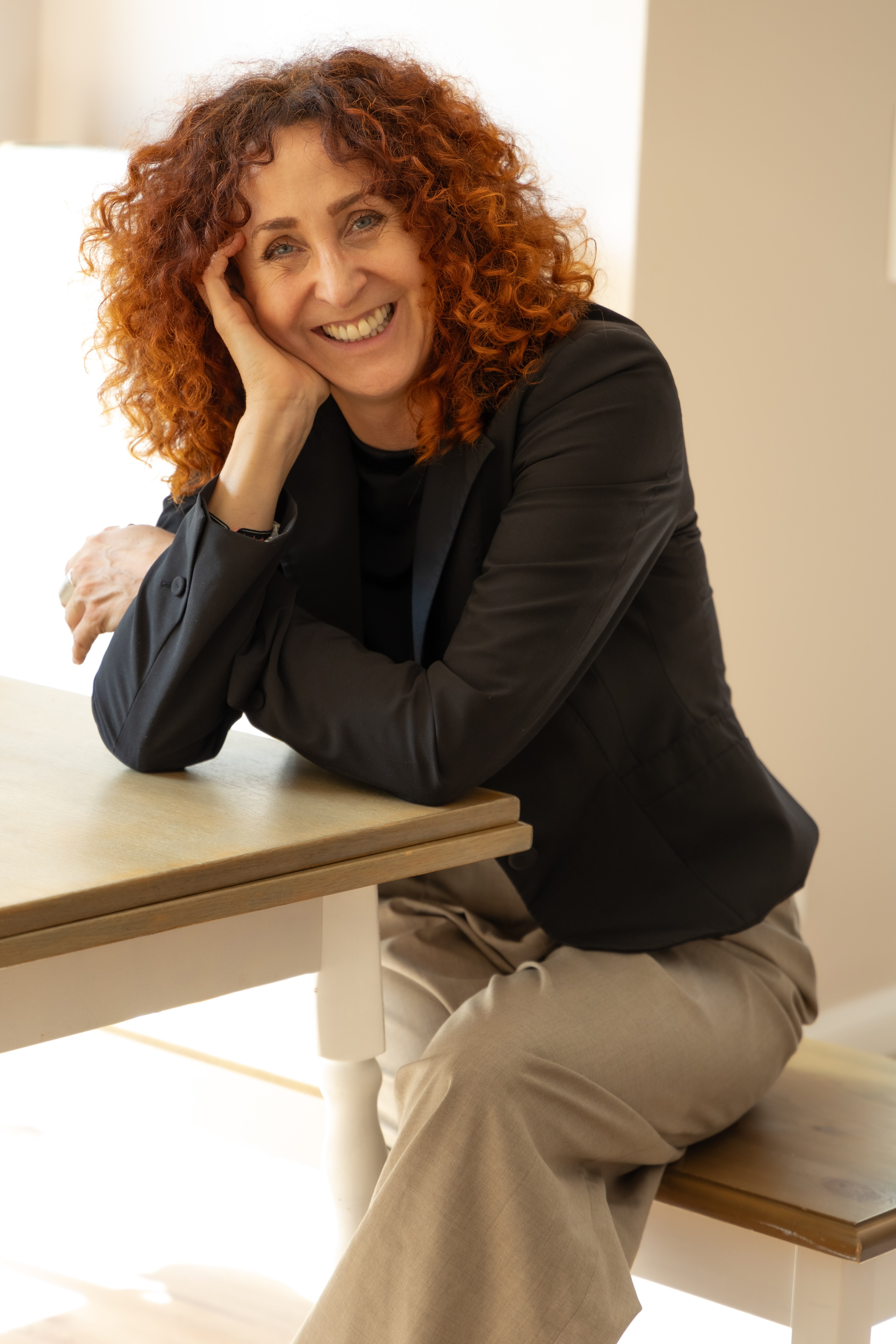 Smiling woman with curly red hair leaning on a wooden table in a bright room.