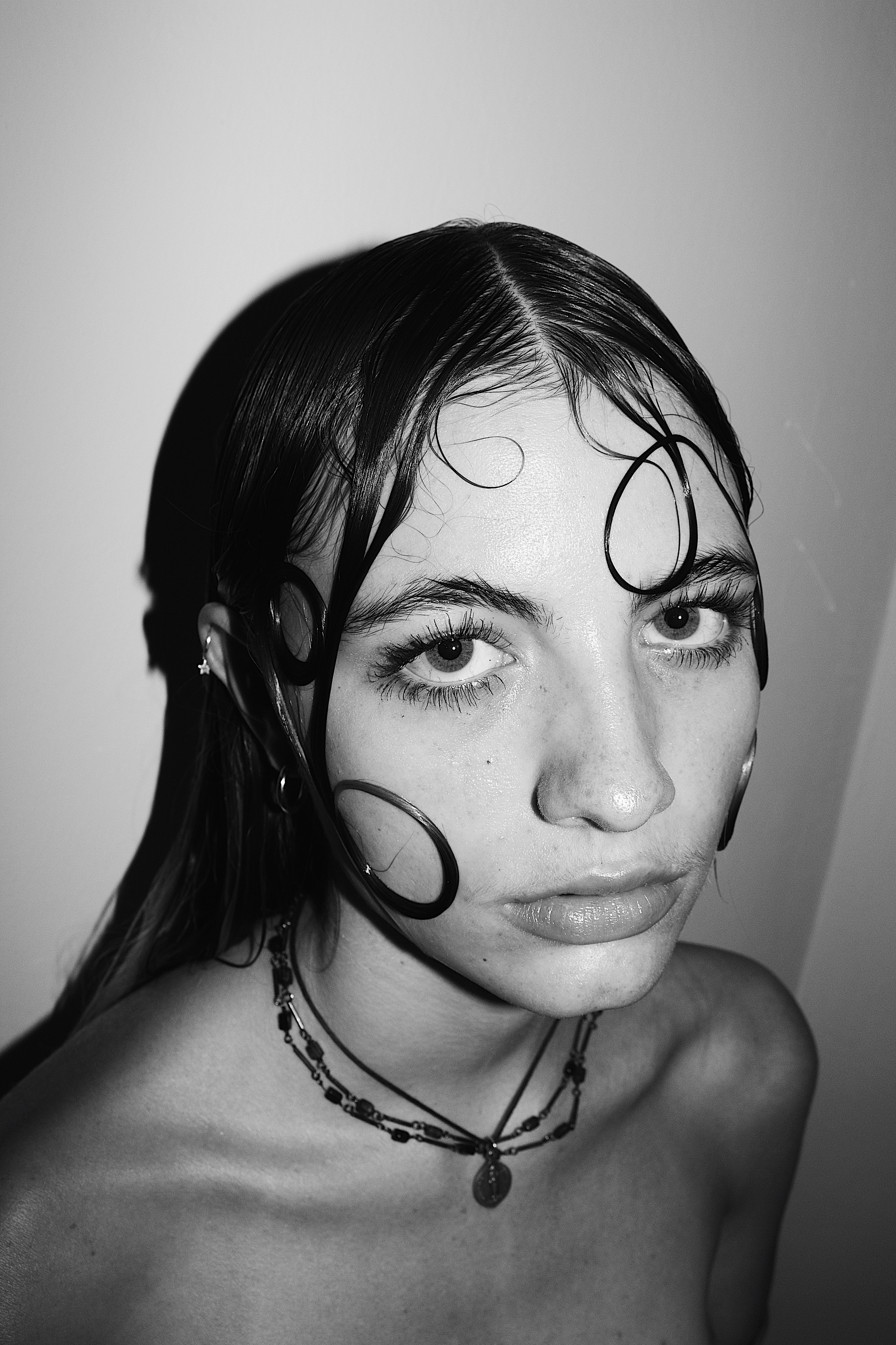 Black and white close-up portrait of a woman with styled wet hair forming loops on her face and wearing layered necklaces.