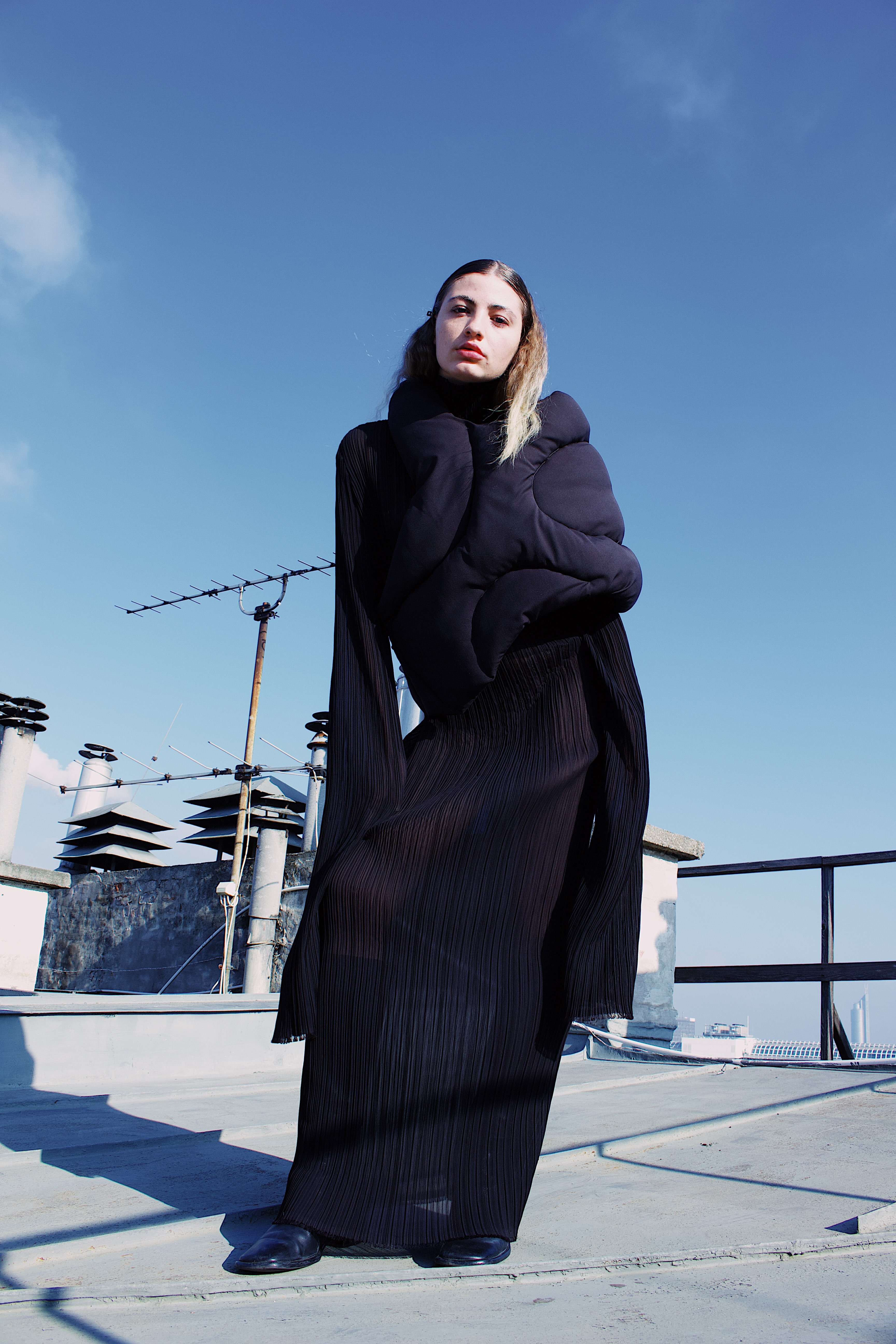 Woman standing on rooftop wearing a long black pleated dress and a large padded black scarf under a clear blue sky.