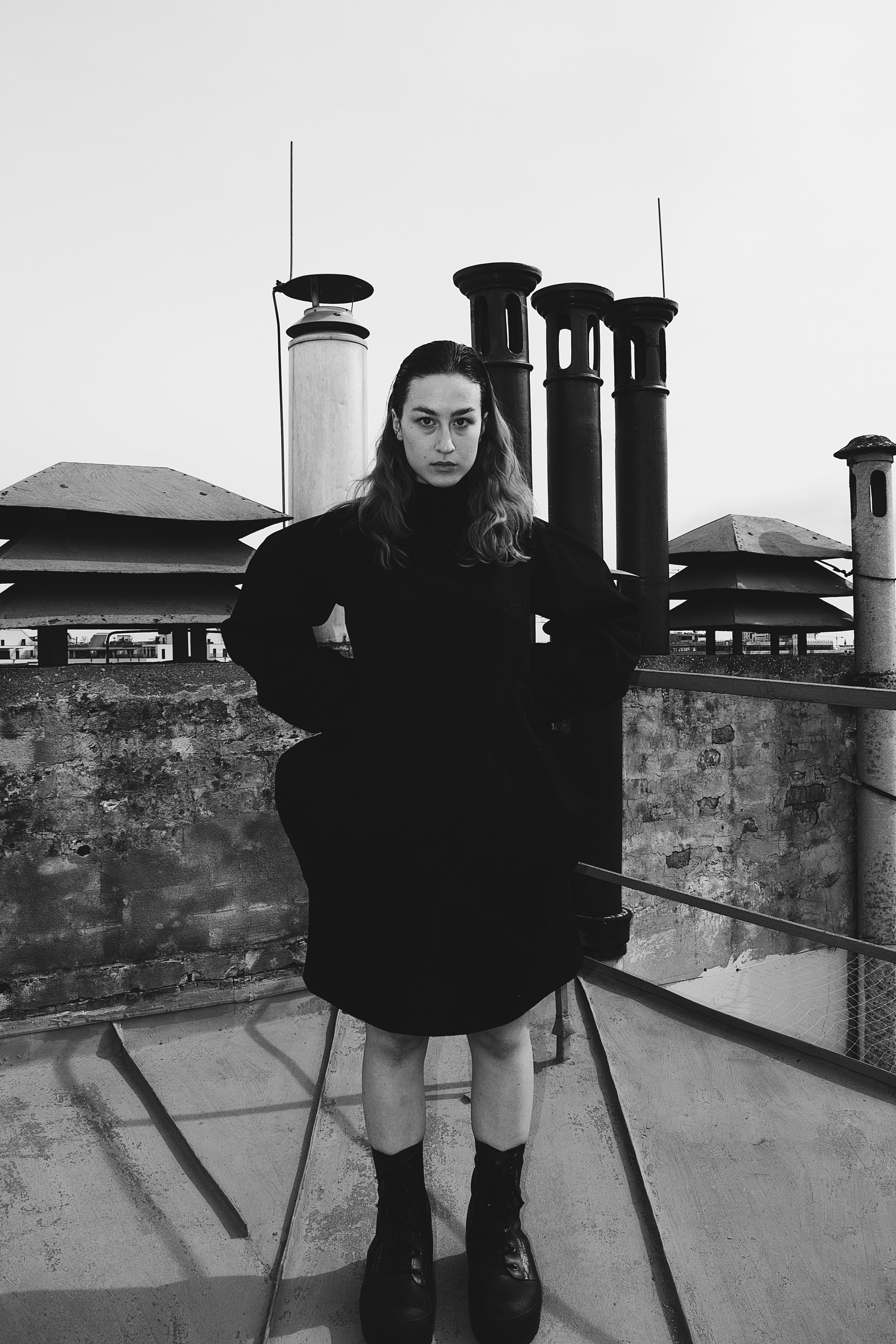 Black and white photo of a serious woman with long hair in a dark dress and boots standing on a rooftop with chimneys behind her.