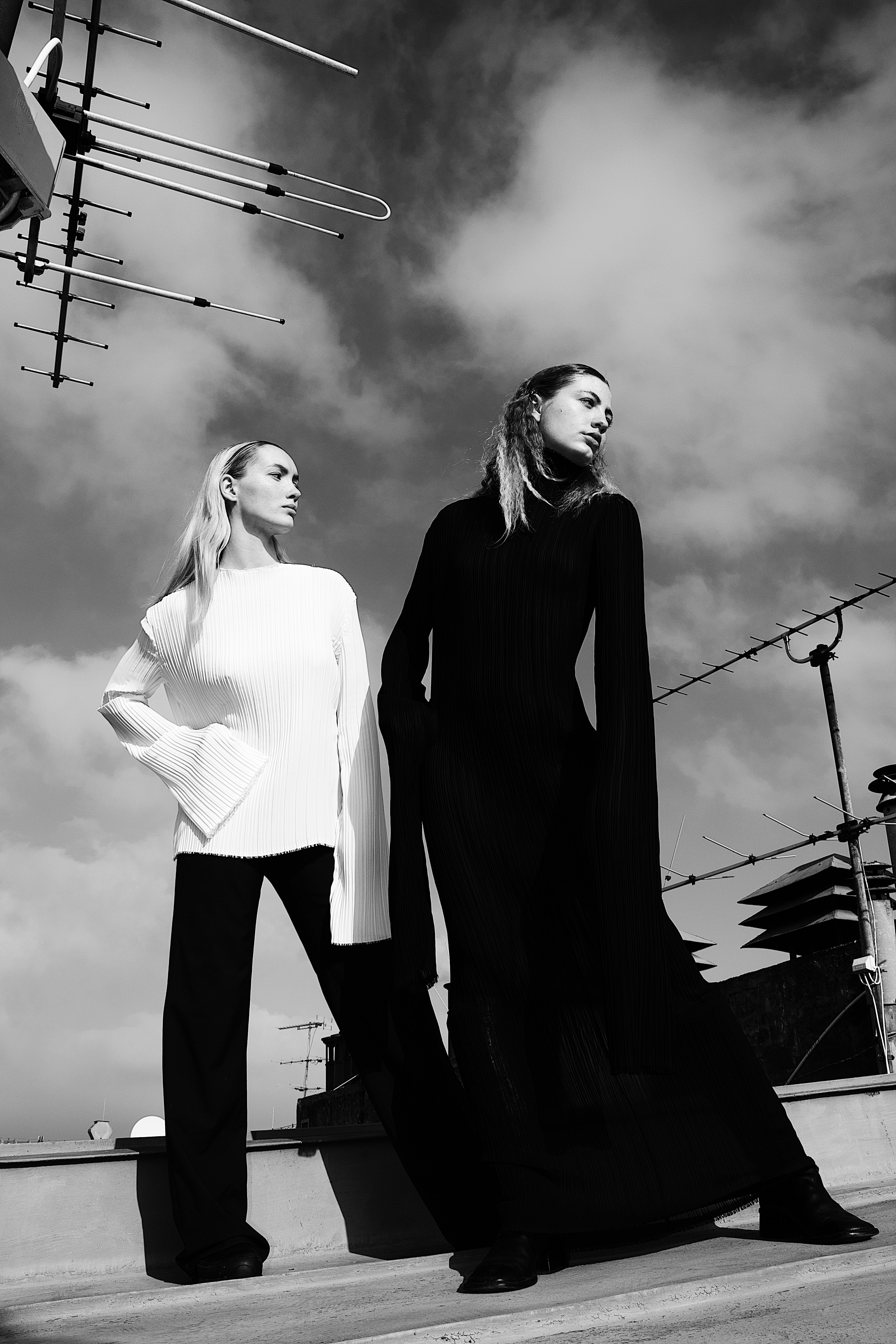 Two women standing on a rooftop wearing contrasting long-sleeved black and white outfits against a partly cloudy sky with TV antennas in the background.
