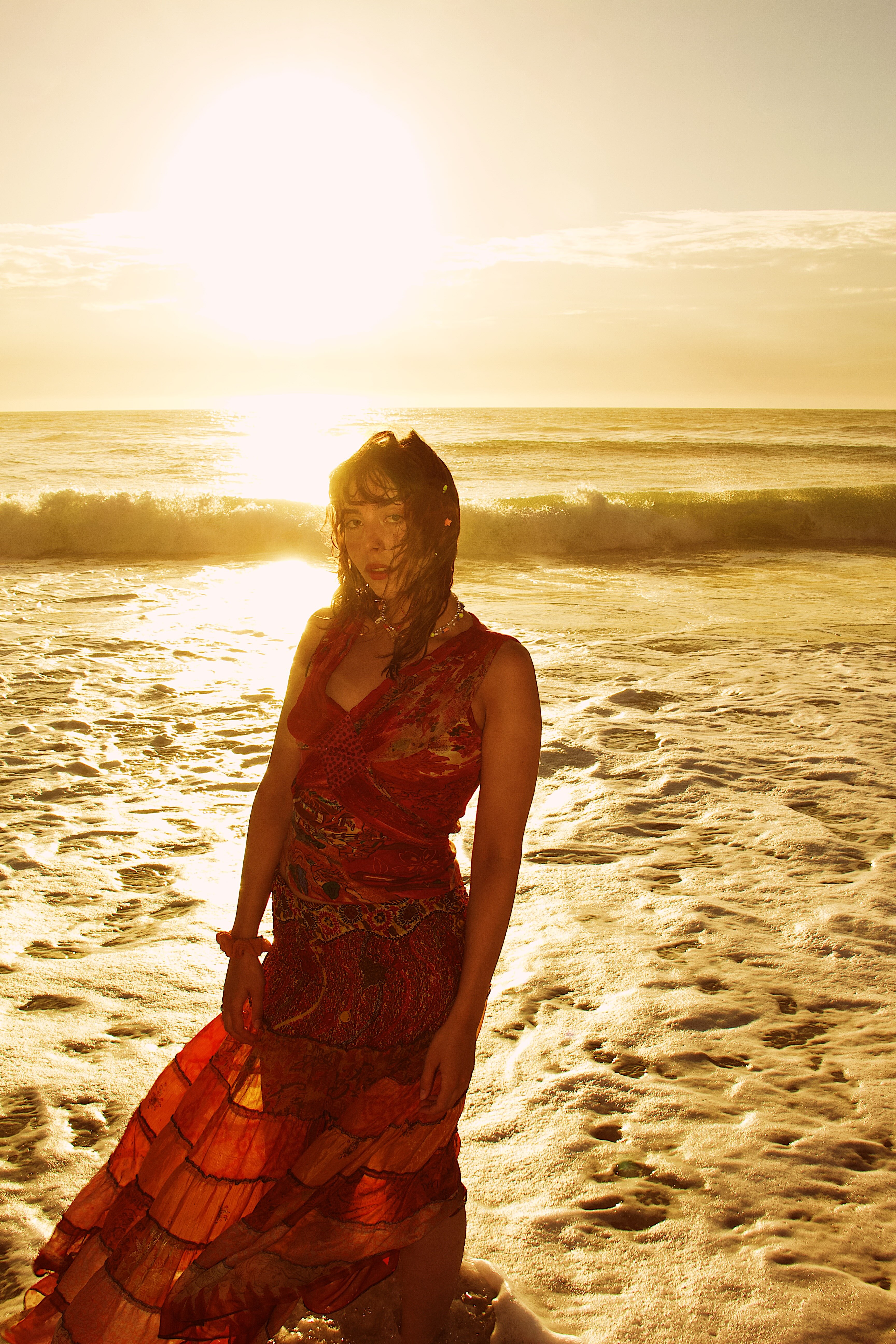 Woman wearing a flowing red dress standing in ocean waves at sunset with sunlight reflecting on the water.