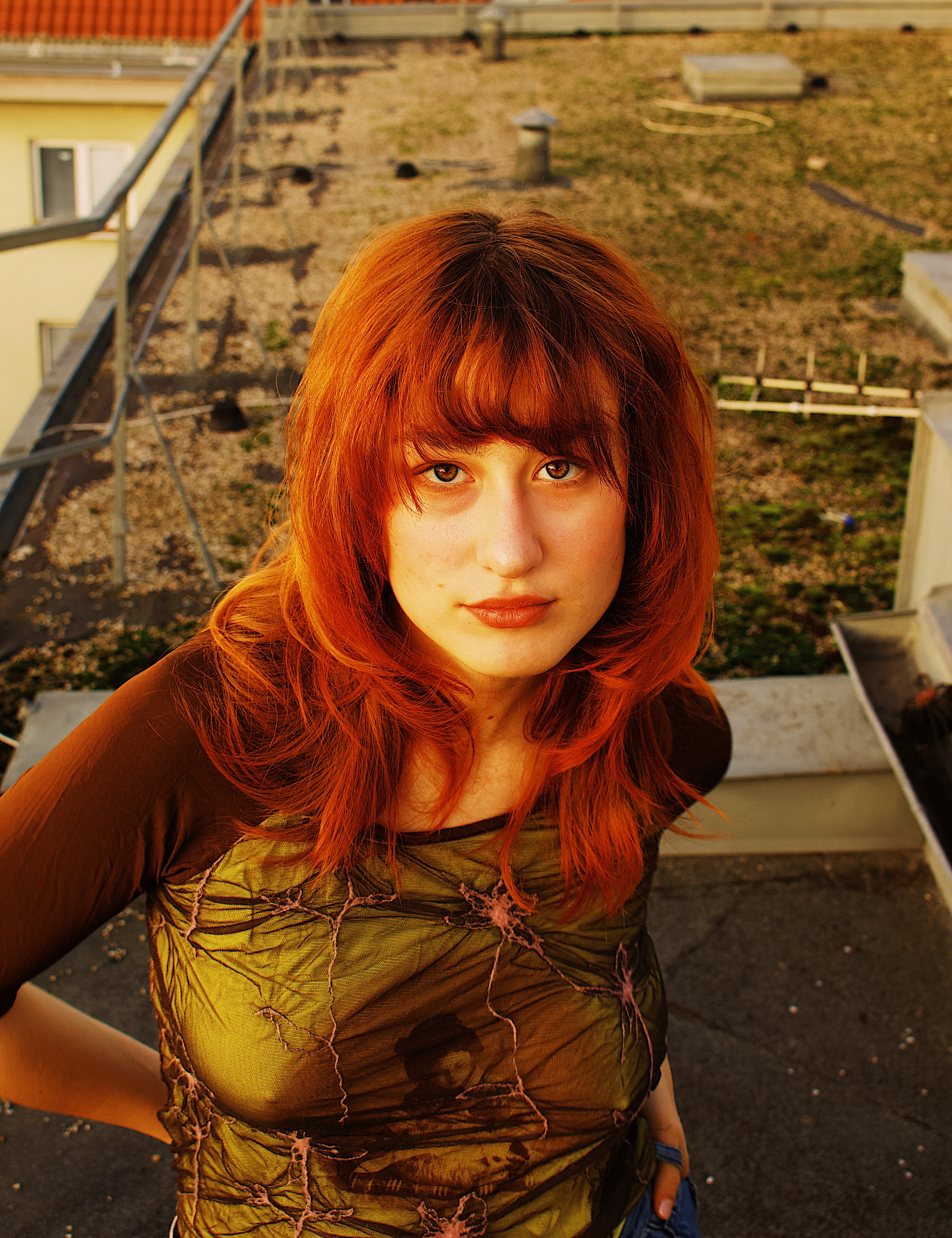 Young woman with bright red hair and bangs wearing a sheer green and brown top, looking directly at the camera outdoors.