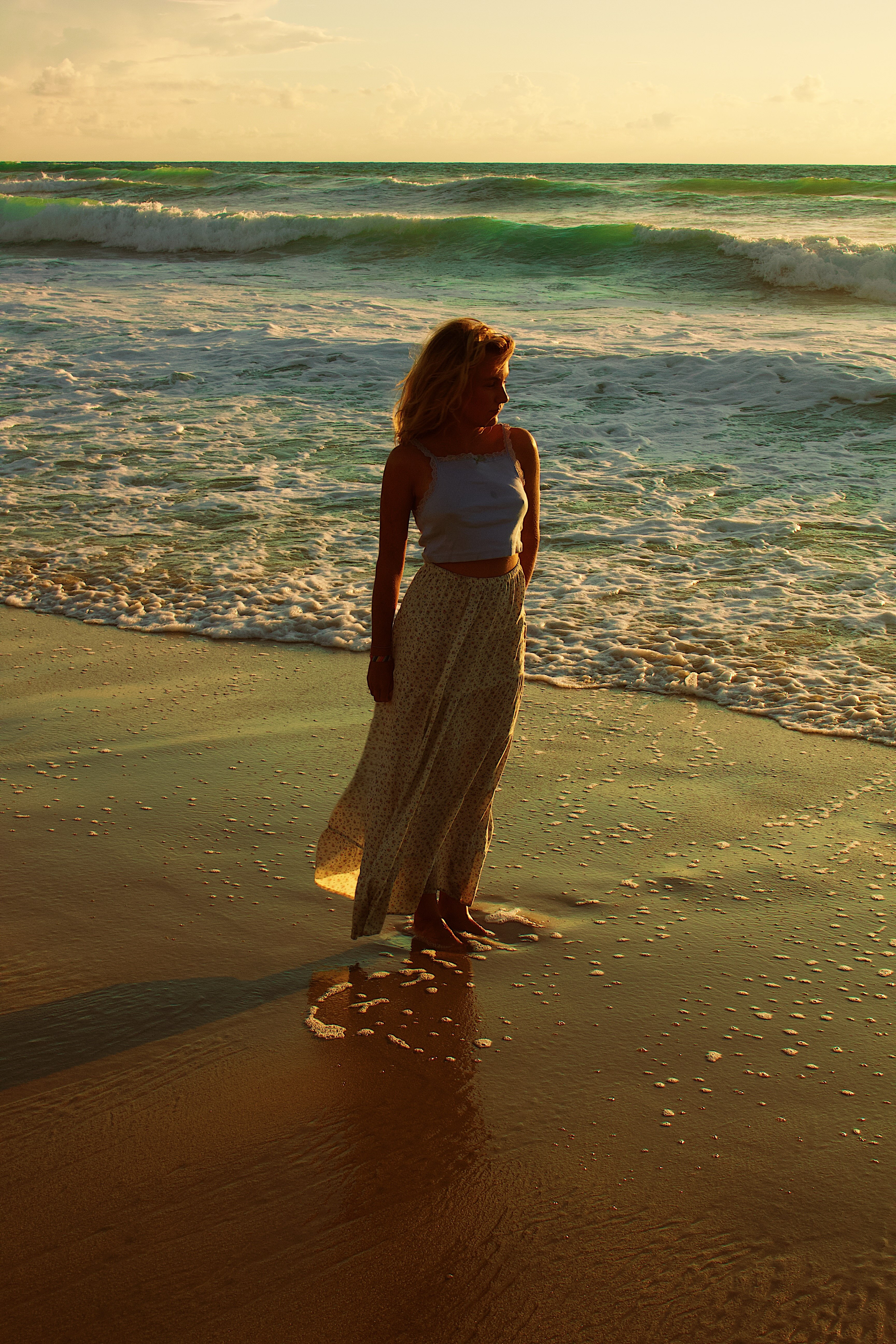 Woman standing barefoot on a sandy beach with waves in the background during sunset.