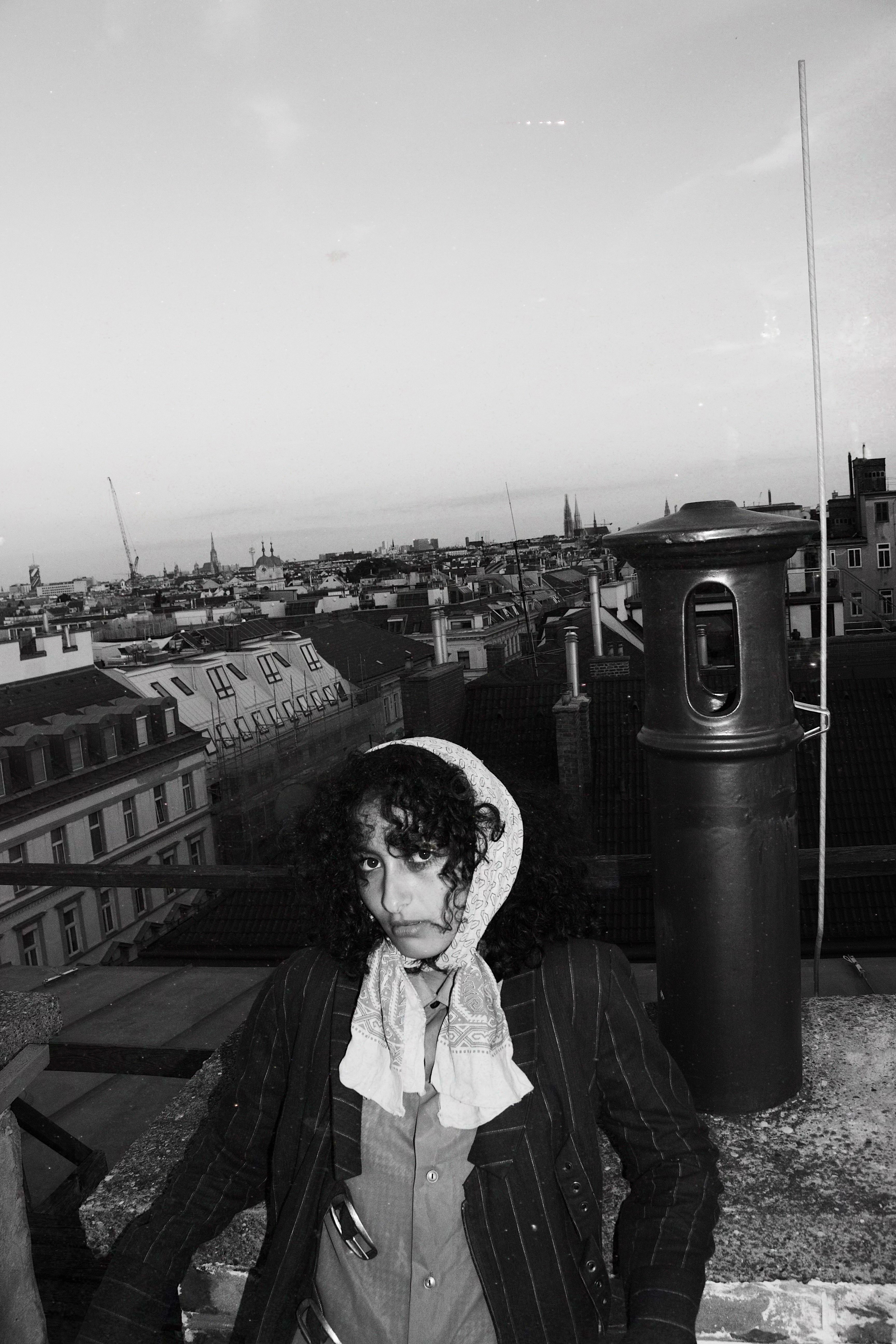 Black and white photo of a woman with curly hair wearing a patterned headscarf and striped jacket, sitting on a rooftop overlooking city buildings.