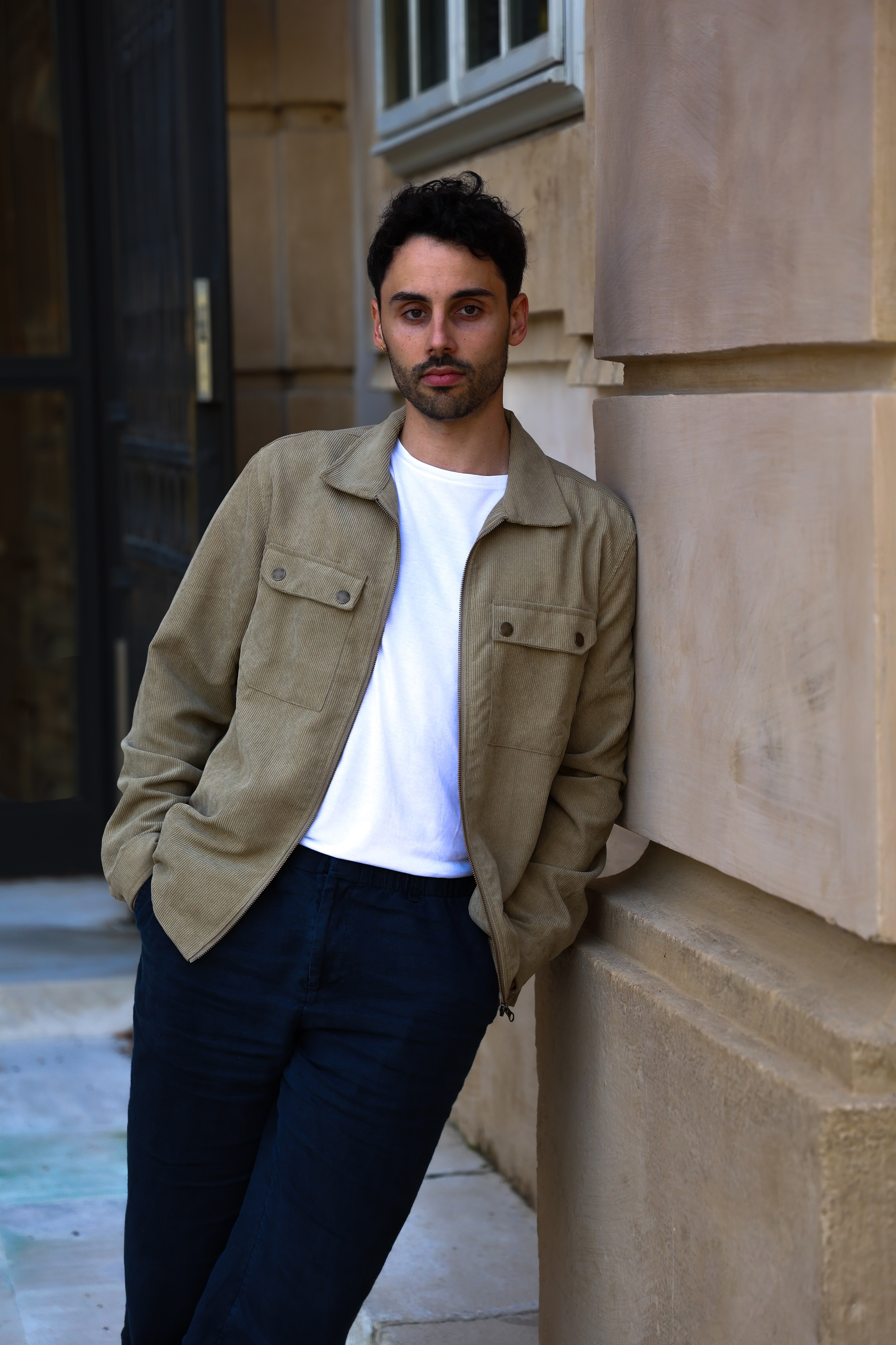 Man with dark hair and light beard wearing a beige jacket, white t-shirt, and dark pants leaning against a beige stone wall outdoors.