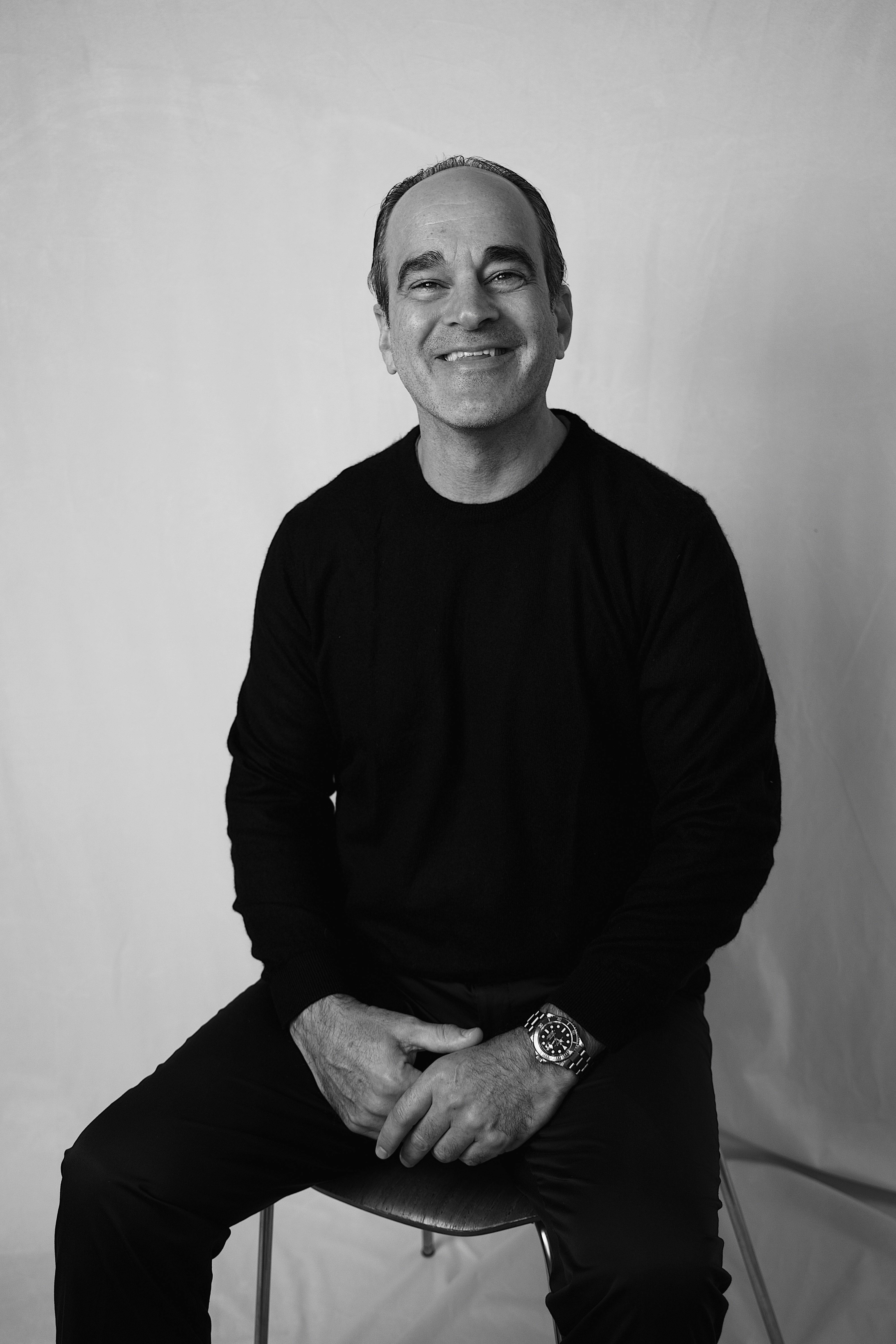 Smiling man in black sweater sitting on a chair against a plain backdrop.