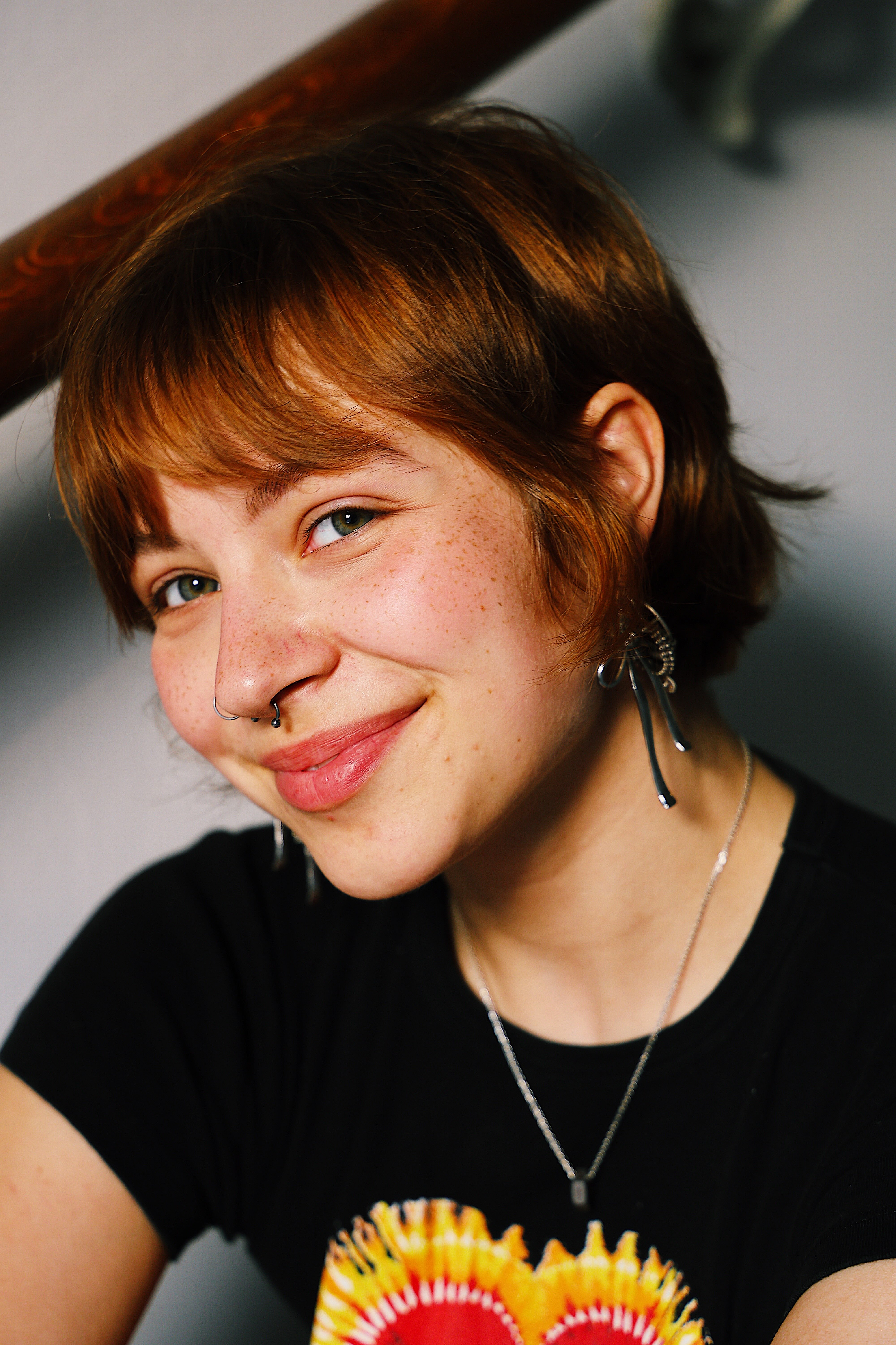 Close-up of a smiling young person with short reddish hair, freckles, septum and nose rings, wearing a black T-shirt and silver necklace.