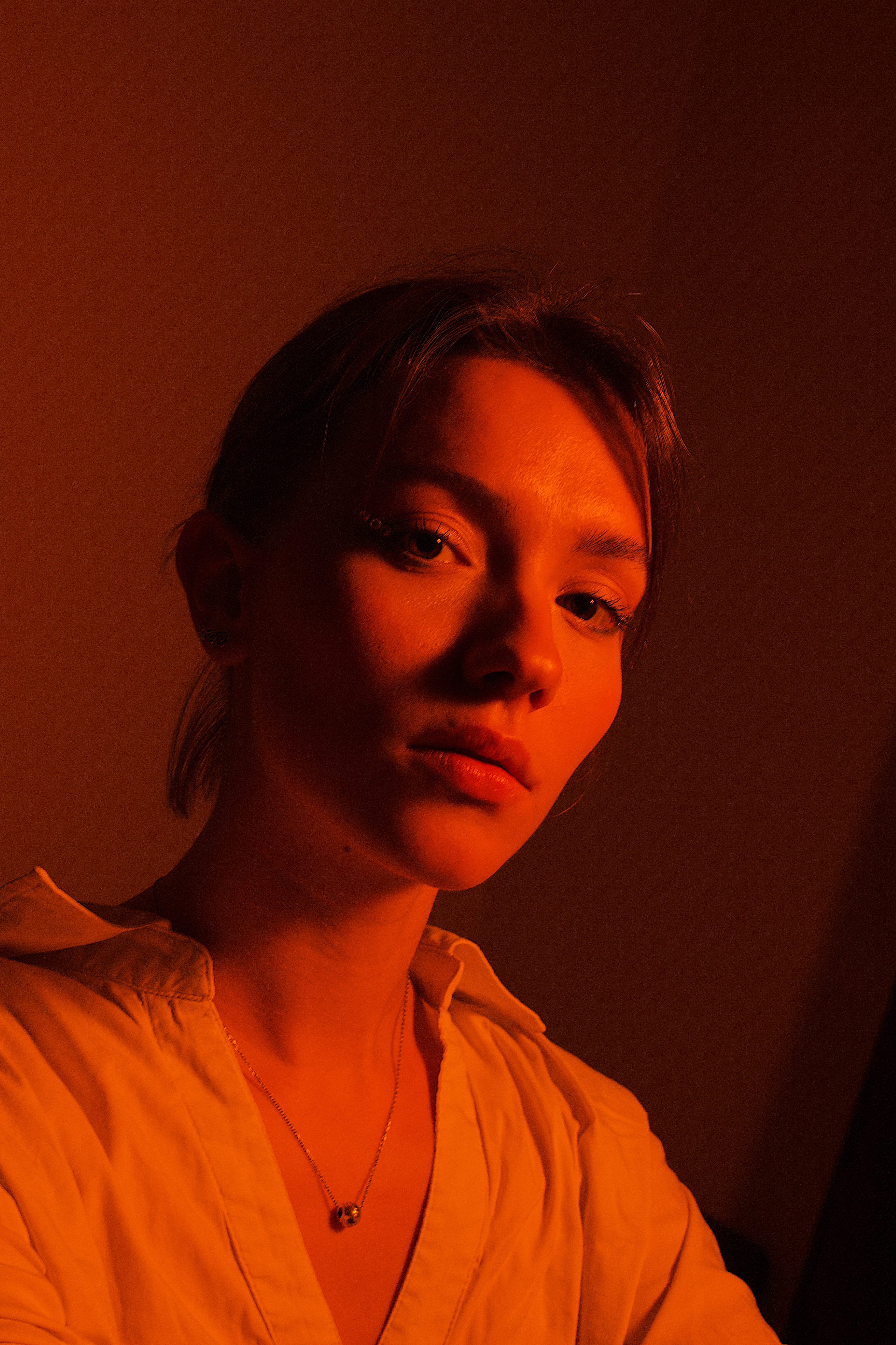 Close-up portrait of a woman with soft orange-red lighting wearing a white shirt and a pendant necklace.