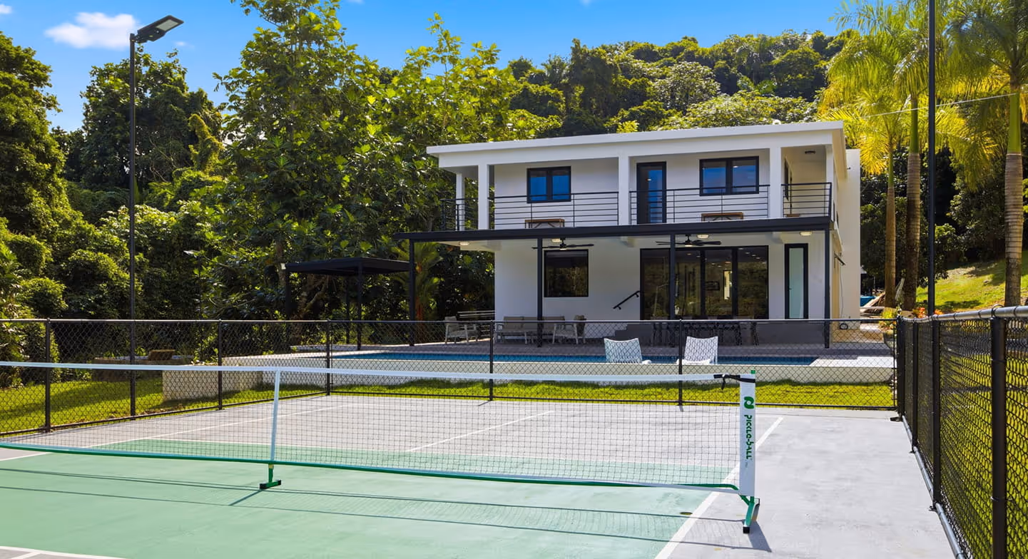 Modern two-story house with a covered patio, pool, and a fenced pickleball court in the foreground surrounded by lush greenery.
