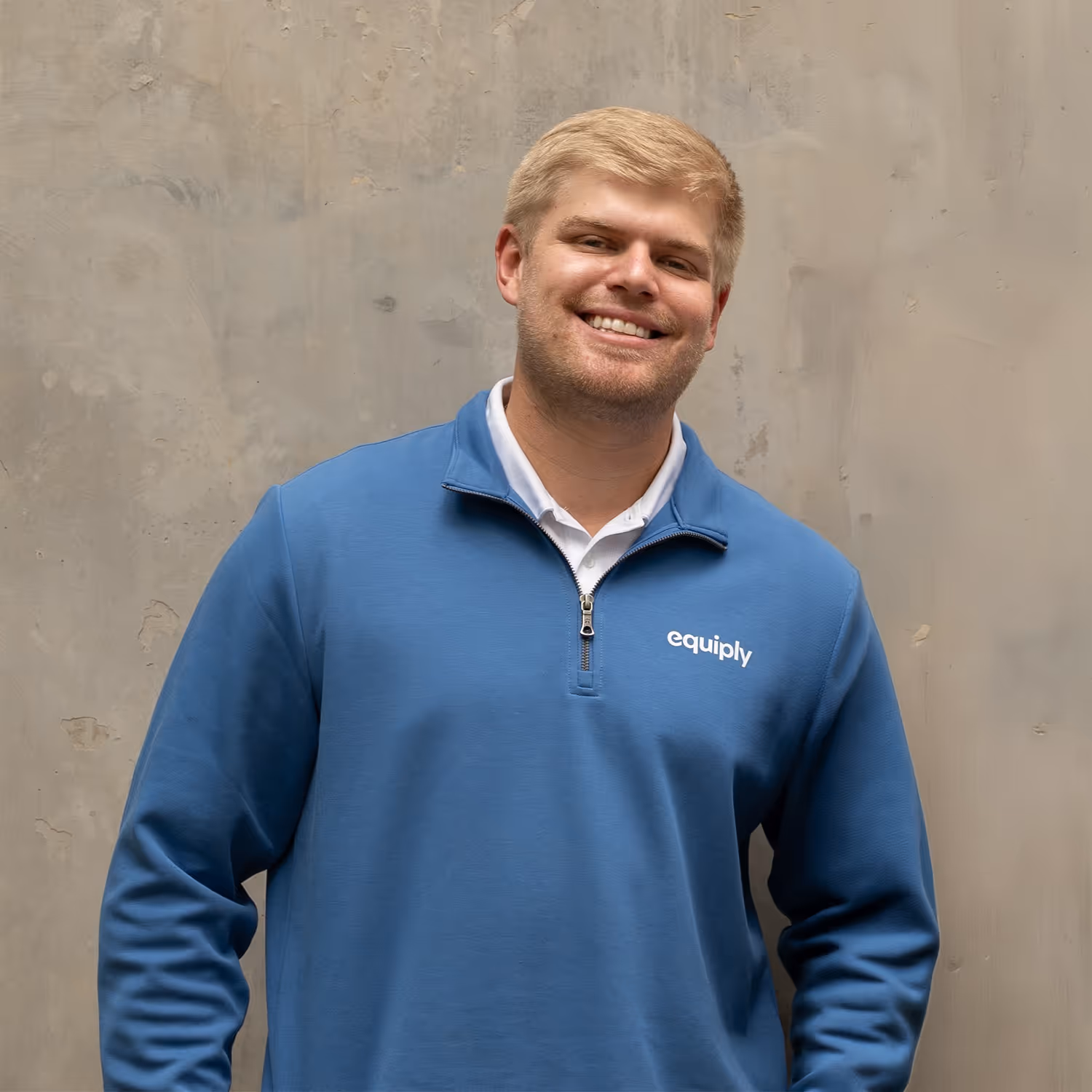 Smiling young man with short blonde hair wearing a blue Equiply quarter-zip pullover and white collared shirt, standing against a textured light gray wall.