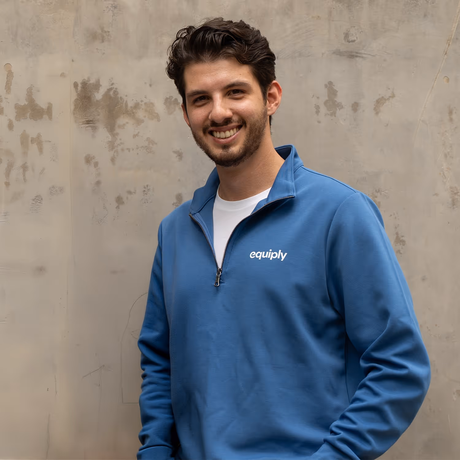 Smiling young man with dark hair and beard wearing a blue Equiply sweatshirt over a white shirt against a textured beige wall.