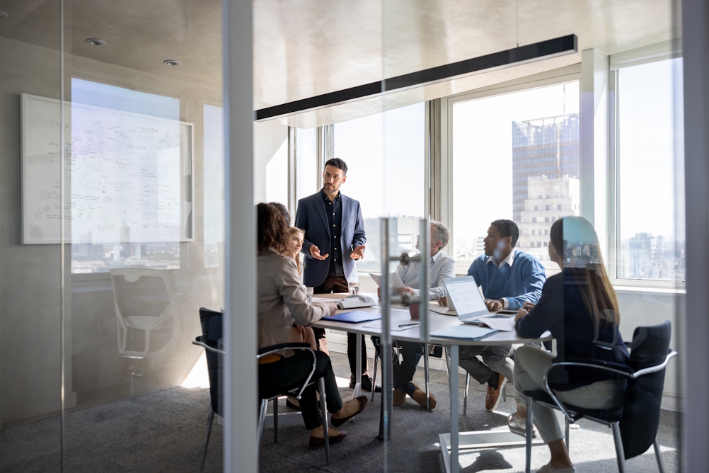 Businessman standing and speaking to colleagues seated around a conference table in a modern office.