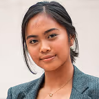 Portrait of a confident young woman with dark hair pulled back, wearing a gray blazer and a heart-shaped necklace.