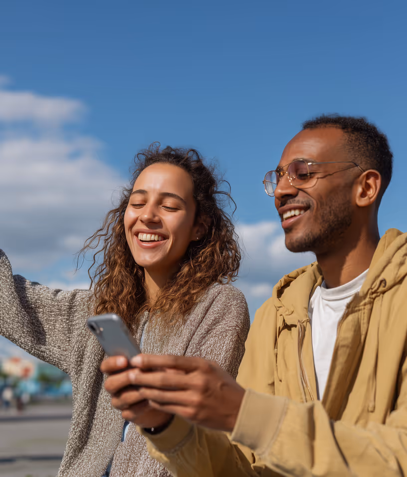 A smiling woman with curly hair and a man wearing glasses looking at a smartphone together outdoors under a blue sky.
