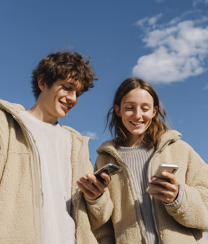 Smiling young man and woman wearing beige fleece jackets looking at their smartphones outdoors against a blue sky.