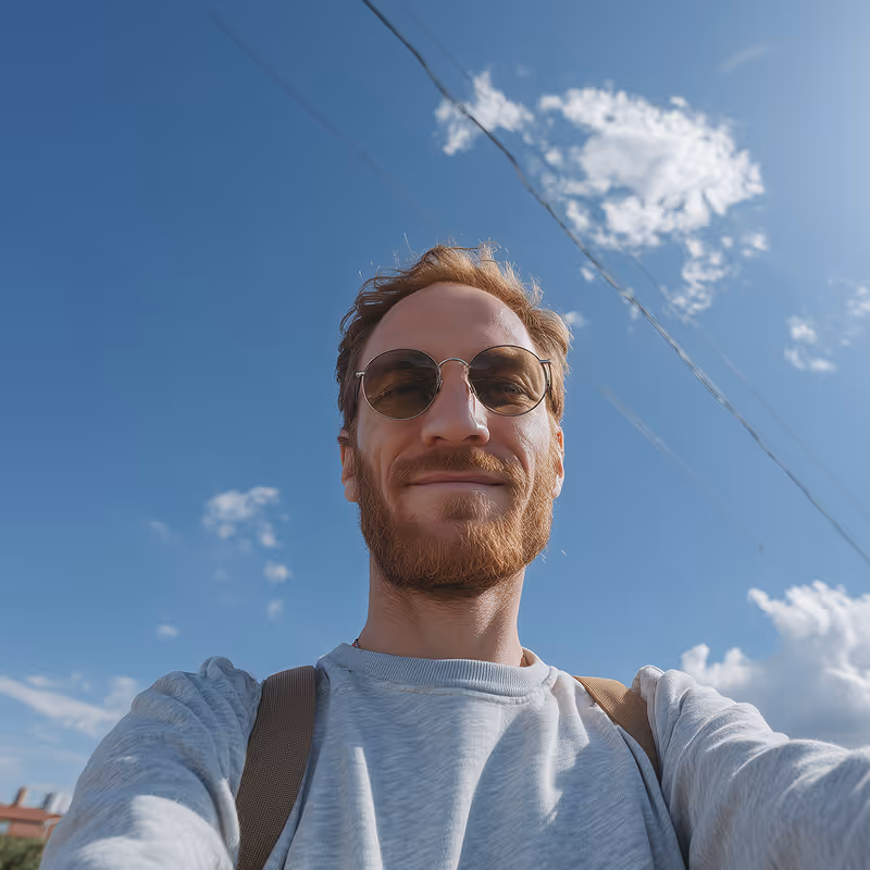 Smiling man with ginger beard and sunglasses taking a selfie outdoors against a blue sky with scattered clouds.