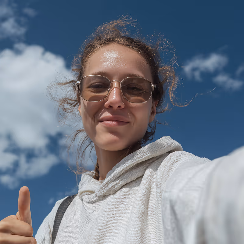 Smiling young woman wearing sunglasses and a white hoodie giving a thumbs up outdoors under a blue sky with clouds.