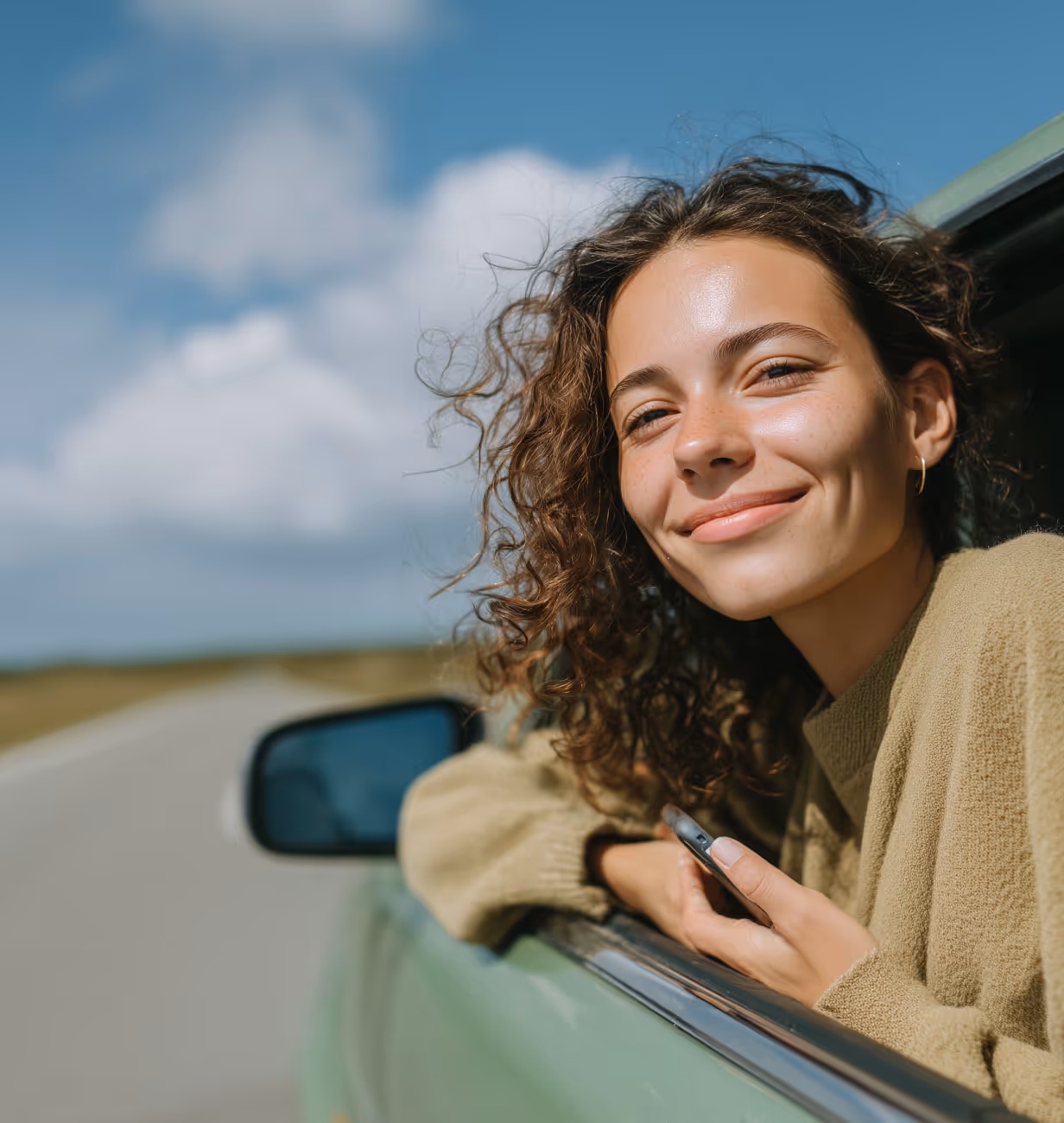 Smiling woman with curly hair leaning out of a car window on a sunny day.