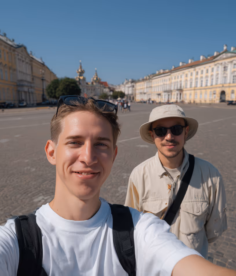 Two young men taking a selfie in a sunny, open square with historic buildings in the background.