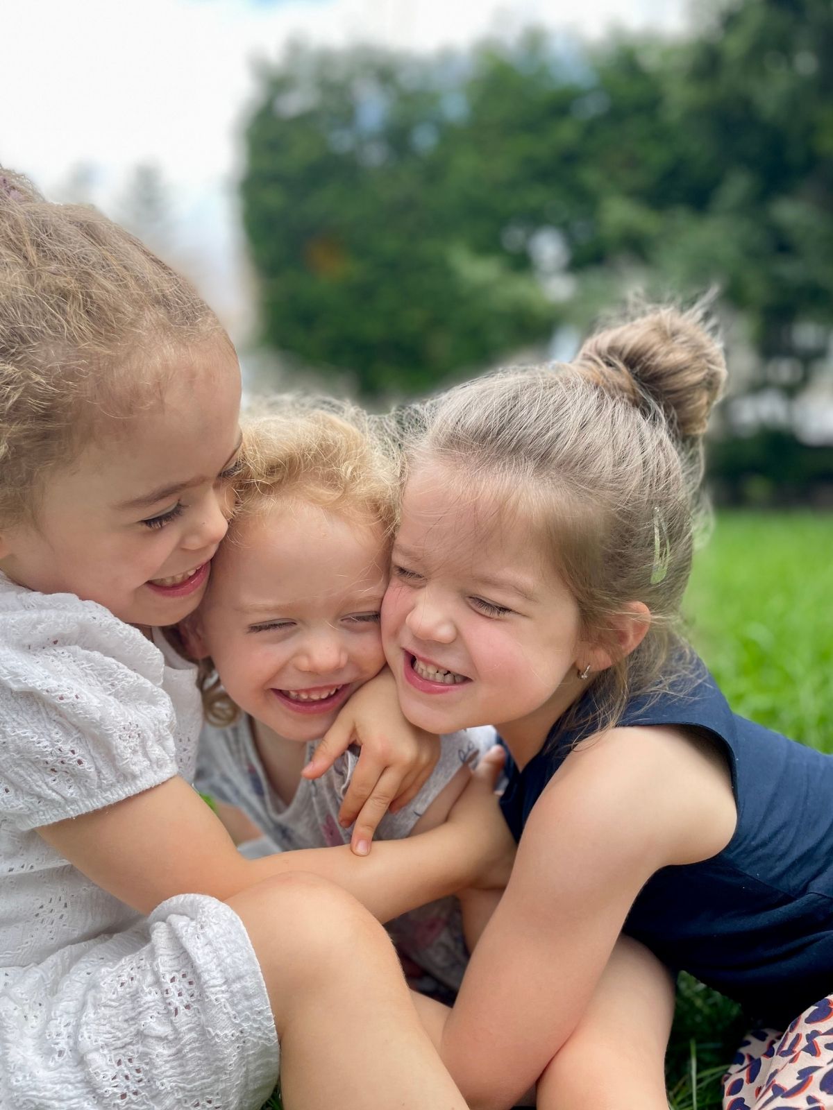 Trois jeunes enfants souriants blottis ensemble dans un moment complice, assis sur l’herbe lors d’une journée ensoleillée.