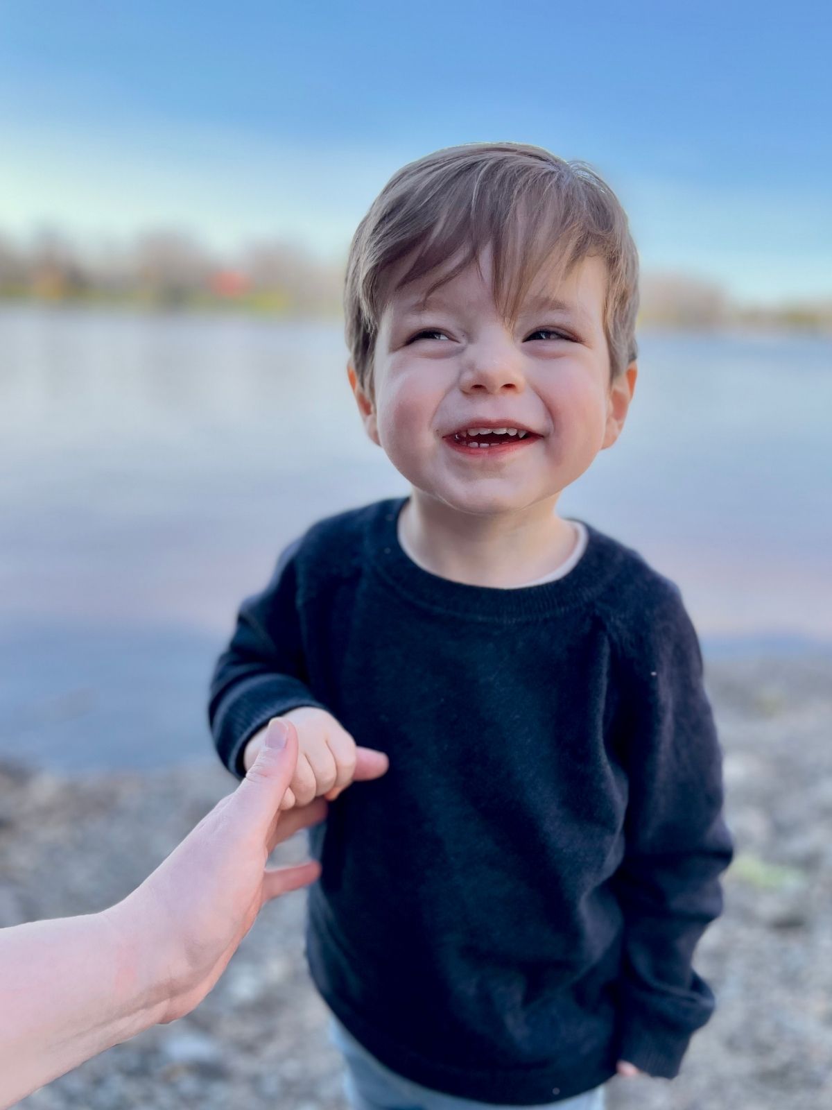 Jeune garçon souriant tenant la main d’un adulte, debout au bord de l’eau sous un ciel dégagé.