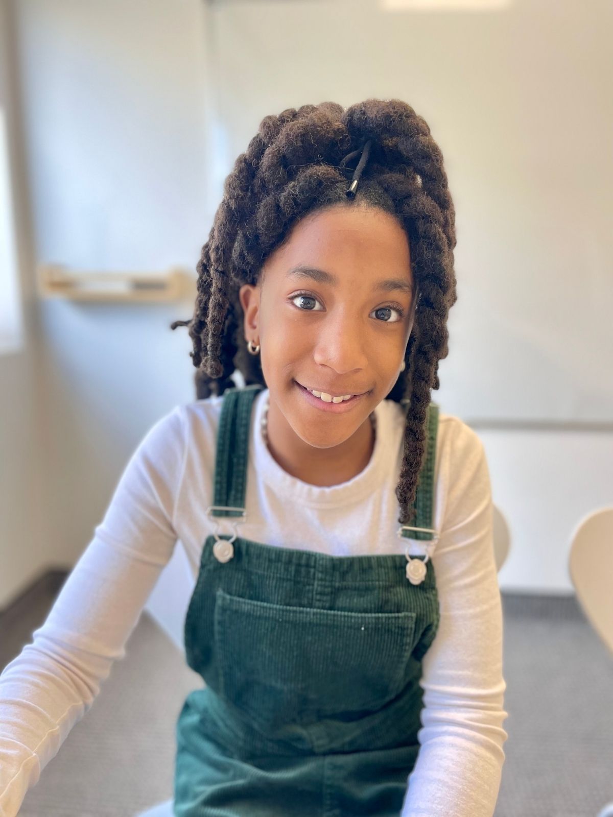 Jeune fille souriante assise en classe, portant une salopette verte et un chandail blanc