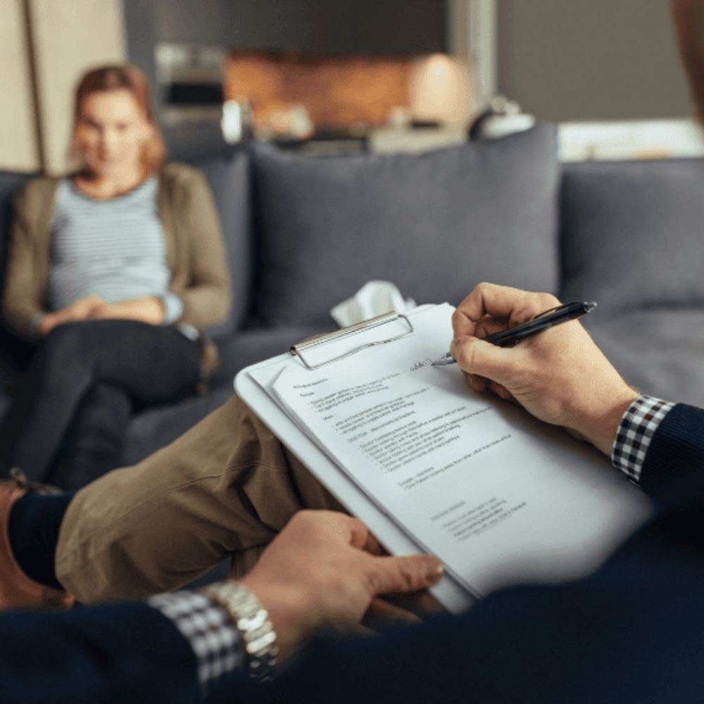 Psychologue prenant des notes pendant qu’une patiente est assise sur un divan en arrière-plan, dans un cadre chaleureux de consultation.
