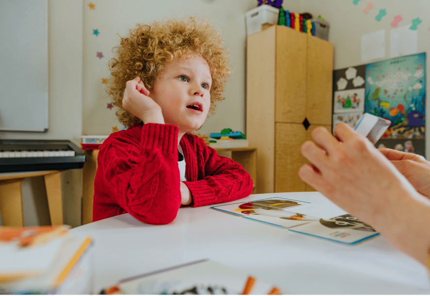 Jeune garçon aux cheveux bouclés blond vénitien, assis à une table en séance d’orthophonie, regardant attentivement une adulte qui tient des cartes d’intervention