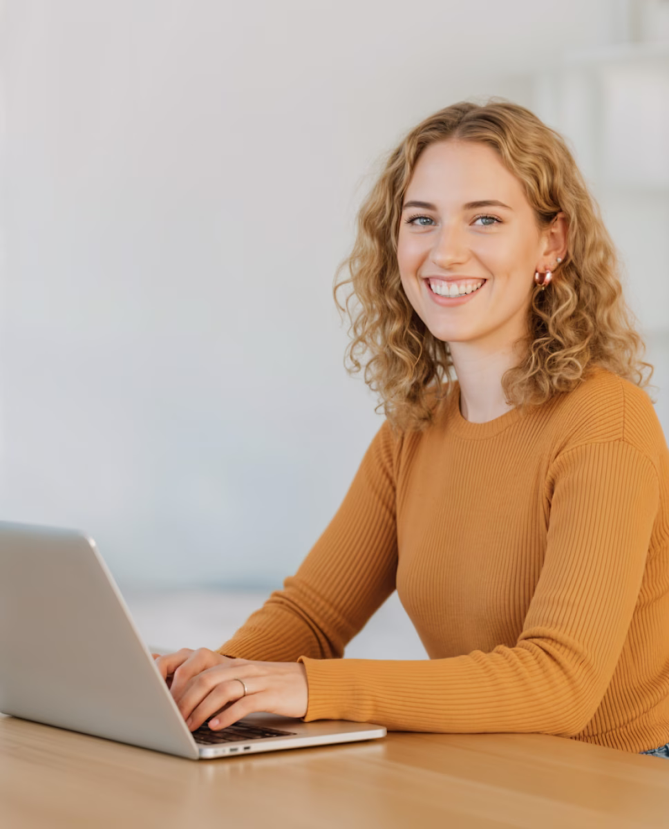 Smiling young woman with curly blonde hair wearing a mustard sweater, typing on a laptop at a wooden table.