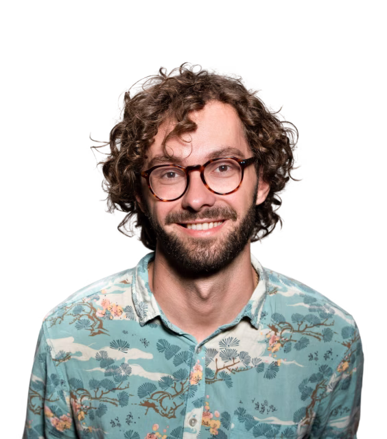 Man with curly hair, beard, and glasses smiling, wearing a turquoise shirt with botanical patterns.