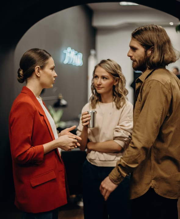 Three young adults having a conversation indoors, with one woman in a red blazer speaking and another woman holding a metal cup while a man listens.