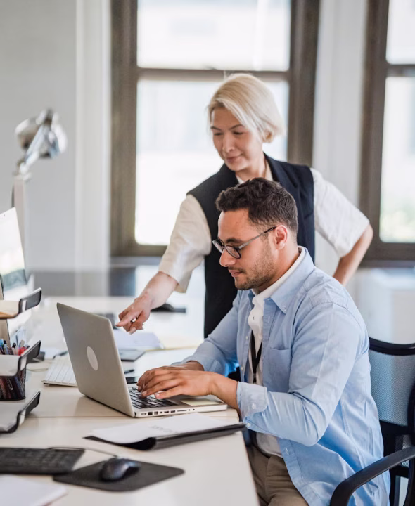 Two coworkers collaborating in an office, with a woman standing and pointing at a laptop screen as a seated man types.