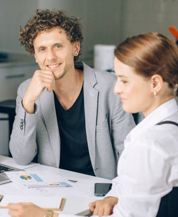 Smiling man in a gray blazer sitting at a desk with charts and a woman in a white shirt nearby.