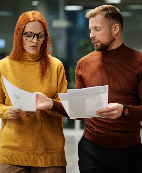 Man and woman wearing sweaters reviewing printed documents together in an office setting.