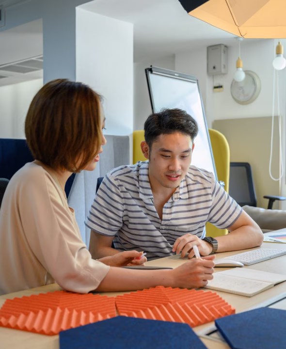 Two young adults collaborating at a table with notebooks and colorful soundproof panels in a modern office space.